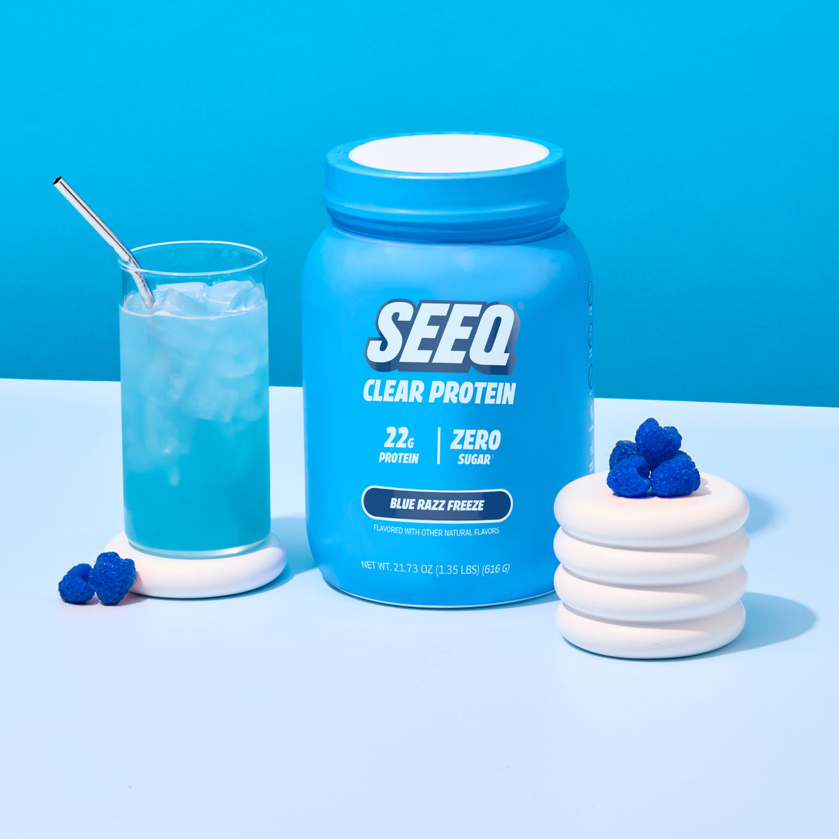 A blue tub of SEEQ Clear Protein powder stands on a white surface against a bright blue background, styled with a glass of blue drink and white gummy candies with blueberries.