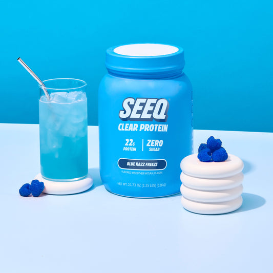 A blue tub of SEEQ Clear Protein powder stands on a white surface against a bright blue background, styled with a glass of blue drink and white gummy candies with blueberries.