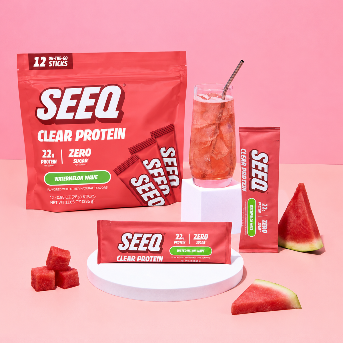 SEEQ Clear Protein watermelon flavor protein powder packaging arranged on a pink studio background, with a large resealable pouch, two single-serve sticks, a glass of pink drink with a straw, and watermelon slices and cubes.