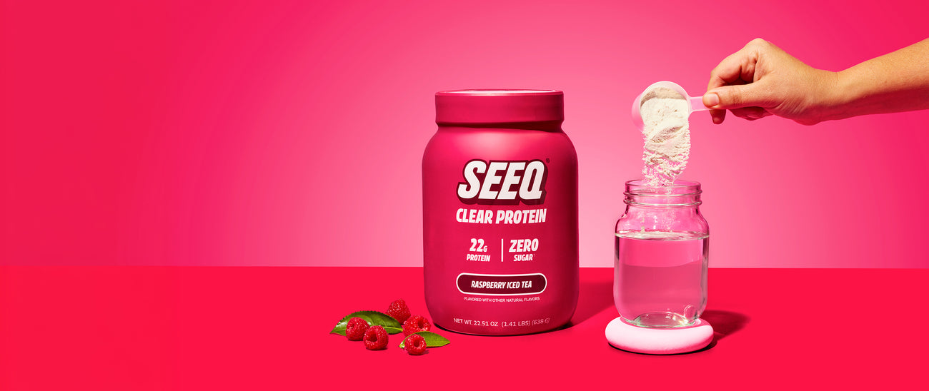 A jar of SEEQ Clear Protein powder in Raspberry Ice Tea flavor, styled on a matching pink background with fresh raspberries at its base.