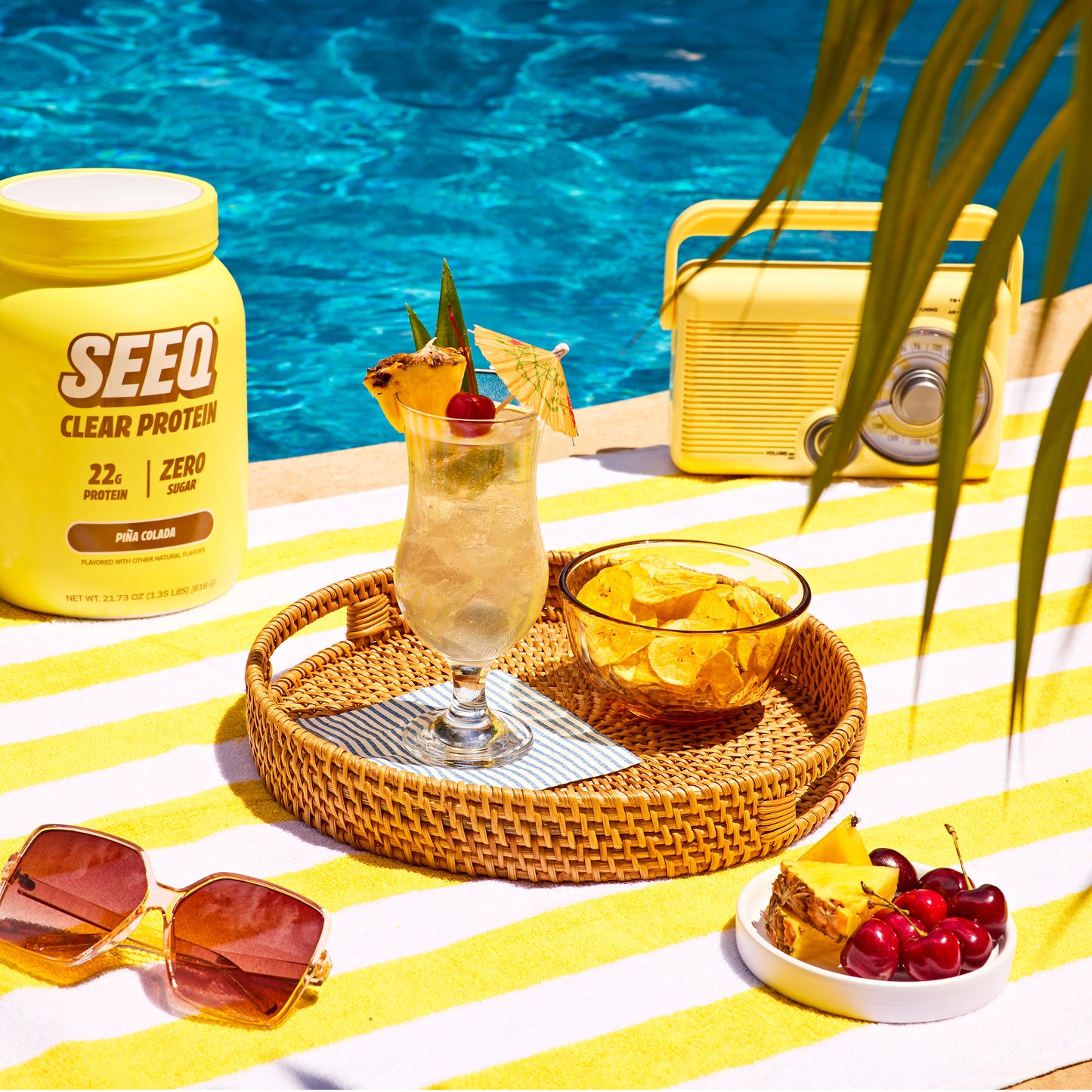 Yellow SEEQ protein container on a striped towel by a pool with a cocktail and fruit.